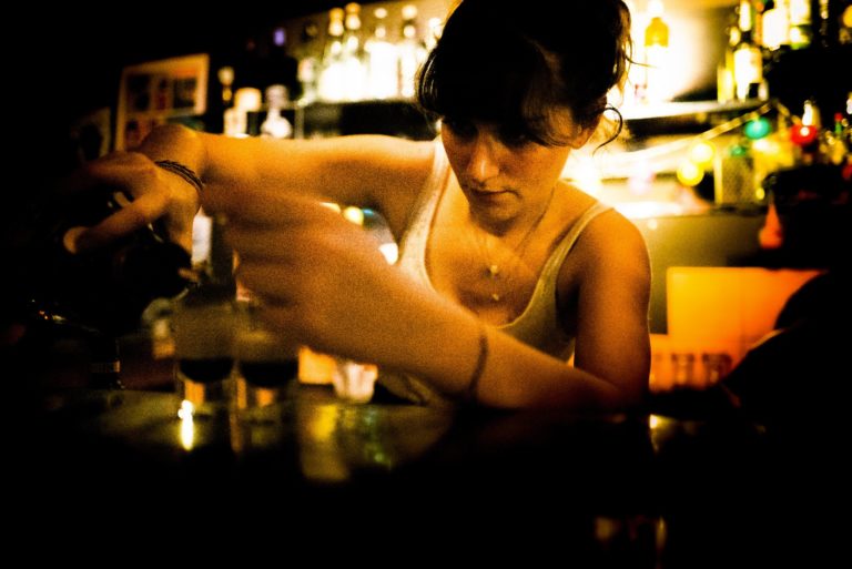 Une barmaid sert au comptoir, les bouteilles éclairées derrière elle